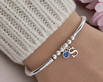 May include: A silver-toned bracelet featuring a series of small metallic beads, a single round blue crystal charm, and a dangling silver-toned letter S charm. The bracelet is constructed with a smooth, curved metal band segment.