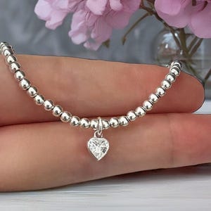 Puede incluir: Una pulsera de cuentas plateadas con un dije en forma de corazón que tiene una piedra transparente facetada en el centro. La pulsera está formada por una serie de pequeñas esferas plateadas, pulidas y reflectantes, unidas en línea.
