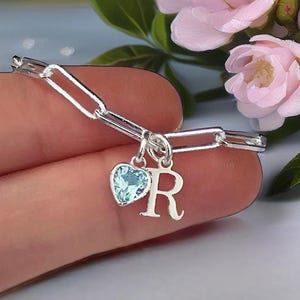Puede incluir: Una pulsera de cadena en tono plateado con dos dijes colgantes: una piedra preciosa azul claro en forma de corazón y una letra R mayúscula. La pulsera está construida con eslabones ovalados alargados y tiene un acabado metálico pulido.