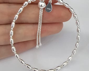 May include: A sterling silver bracelet featuring a series of polished, elongated oval beads interspersed with small round spheres. The bracelet includes an adjustable sliding clasp mechanism with two dangling chains and a teardrop tag marked S925.