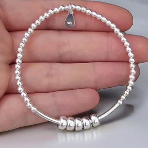 Puede incluir: Una pulsera elástica de plata de ley compuesta por pequeñas cuentas redondas y cinco cuentas cilíndricas más grandes. Un pequeño dije en forma de lágrima cuelga en la parte superior, grabado con el texto 925, indicando la pureza de la plata.