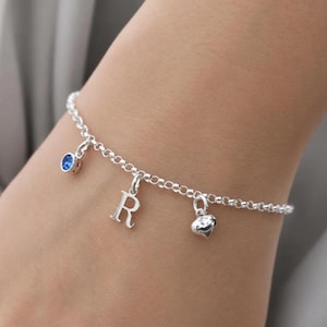 Peut inclure: Un bracelet en chaîne argentée avec trois breloques pendantes : une petite pierre précieuse bleue circulaire, une lettre majuscule R et un cœur lisse. Le bracelet est composé de maillons ronds avec une finition métallique polie.