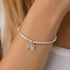 May include: A silver beaded stretch bracelet featuring a dangling charm of the letter K and a small, pink, teardrop-shaped gemstone. The bracelet is constructed from a series of uniform, polished silver-toned beads strung on a flexible cord.