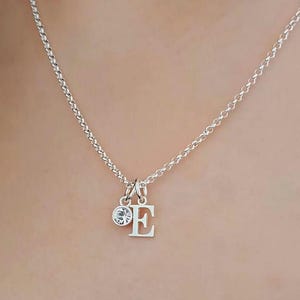 Puede incluir: Collar de plata con una cadena delicada. Presenta una pequeña gema transparente y un colgante de plata con la letra "E". El colgante cuelga de la cadena, creando un diseño sencillo y elegante.