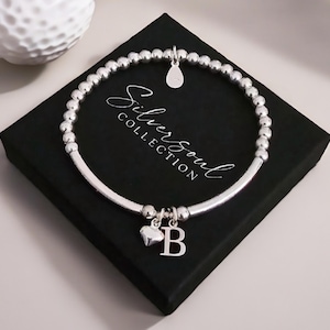 Peut inclure: Un bracelet élastique composé de perles argentées, agrémenté d'une petite breloque en forme de goutte, d'un cœur bombé et d'une lettre B. Le bijou est présenté sur une boîte carrée noire portant l'inscription Silver Soul Collection en blanc.