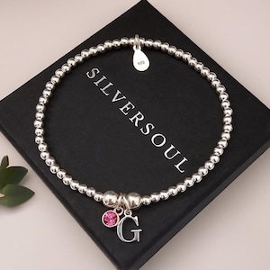 Könnte beinhalten: Ein elastisches Armband aus silberfarbenen Perlen mit einem kleinen tropfenförmigen Anhänger mit 925-Prägung, einem runden rosa Kristallanhänger und einem silbernen Buchstaben-Anhänger G. Das Armband liegt auf einer schwarzen Box mit Aufschrift SILVERSOUL.