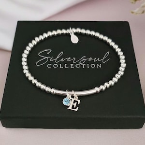 May include: A silver beaded stretch bracelet featuring a small dangling charm with the letter E and a round light blue crystal. The bracelet is displayed on a black square box with the text Silversoul COLLECTION printed in white script and font.