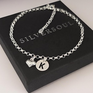 Op de afbeelding: Een zilveren kettingarmband met een kleine gepolijste hartvormige bedel en een ronde bedel gegraveerd met de letter K. Het armbandje ligt op een zwarte doos met de tekst SILVERSOUL in witte letters op de bovenzijde gedrukt.
