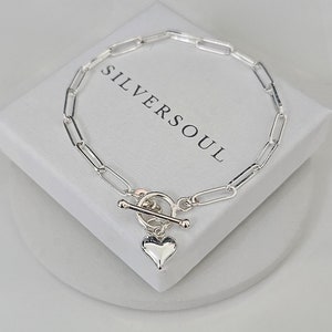 Puede incluir: Un brazalete de cadena de plata con un dije de coraz&oacute;n y un cierre de palanca. El brazalete est&aacute; sobre una caja blanca con el texto "SILVERSOUL" impreso en ella.
