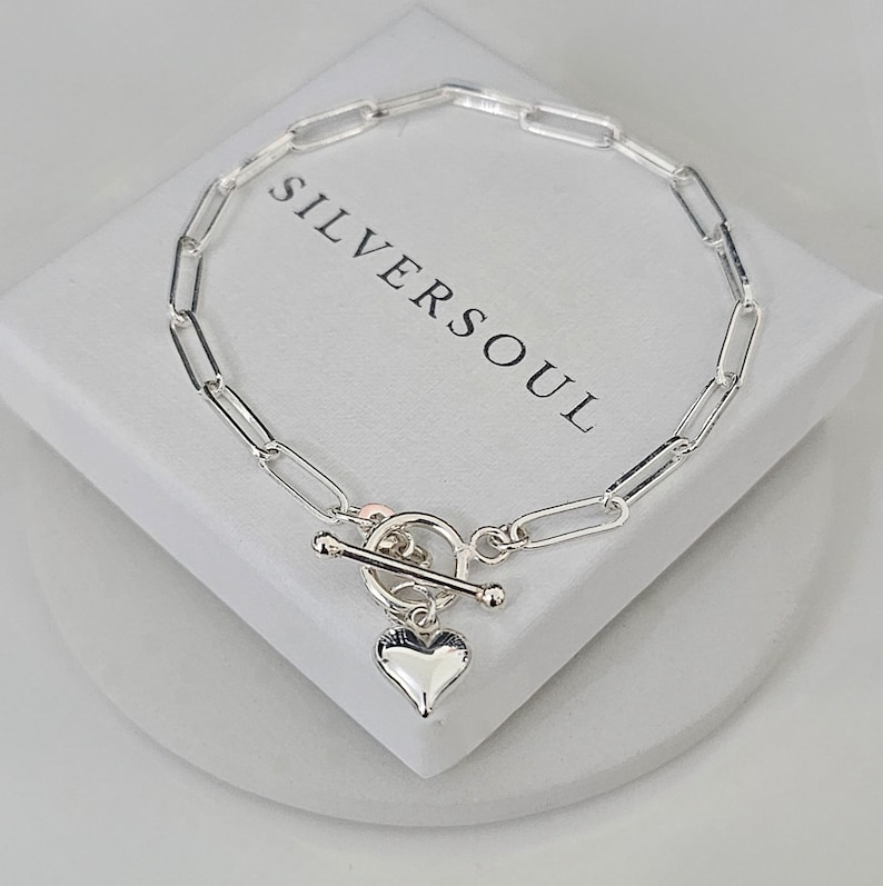 Puede incluir: Un brazalete de cadena de plata con un dije de coraz&oacute;n y un cierre de palanca. El brazalete est&aacute; sobre una caja blanca con el texto "SILVERSOUL" impreso en ella.