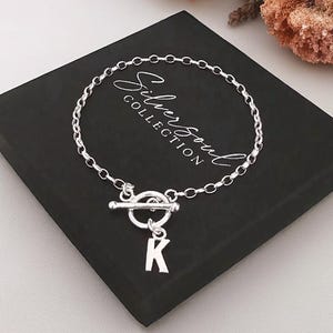 May include: A silver chain bracelet featuring a toggle clasp and a hanging letter K charm. The bracelet is displayed on a black square box with the white text Silver Soul Collection printed on the top surface of the box.