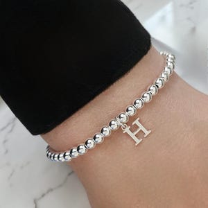 Può includere: Braccialetto con perline d'argento con un ciondolo a forma di lettera "H". Il braccialetto è composto da piccole perline rotonde e lucenti d'argento. Il ciondolo è una lettera maiuscola "H" in un carattere semplice ed elegante.