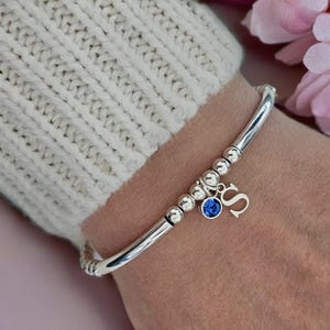 May include: A silver-toned bangle bracelet featuring a series of small metallic beads, a circular charm with a vibrant blue stone, and a dangling letter S charm. The bracelet is constructed with a smooth, polished metal band and a flexible design.