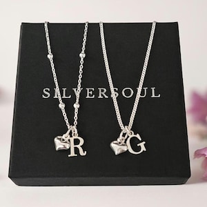 Può includere: Due collane in argento presentate su una scatola nera con il testo SILVERSOUL. Una collana presenta una catenina sottile con piccole perline, un ciondolo a forma di cuore e la lettera R. L'altra collana ha una catena standard, un cuore e la lettera G.