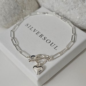 Puede incluir: Un brazalete de cadena de plata con un dije de coraz&oacute;n y un cierre de palanca. El brazalete est&aacute; sobre una caja blanca con el texto "SILVERSOUL" impreso en ella.