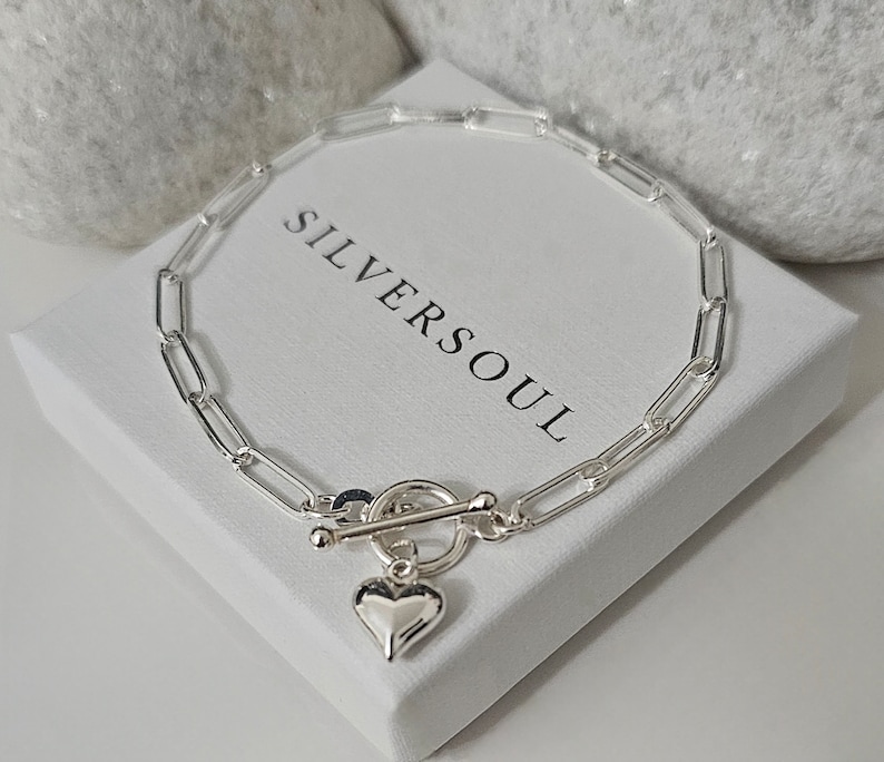 Puede incluir: Un brazalete de cadena de plata con un dije de coraz&oacute;n y un cierre de palanca. El brazalete est&aacute; sobre una caja blanca con el texto "SILVERSOUL" impreso en ella.