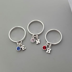 May include: Three silver-toned rings, each featuring a dangling charm set with a round coloured gemstone and a silver-toned letter charm. The first ring has a blue stone and the letter E, the second a pink stone and letter N, and the third a red stone and letter H.
