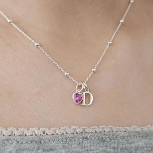 Puede incluir: Un collar de plata con una delicada cadena con pequeños detalles de bolas. El colgante incluye un dije en forma de corazón rosa y la letra "D" plateada. El collar se muestra sobre un fondo neutro.