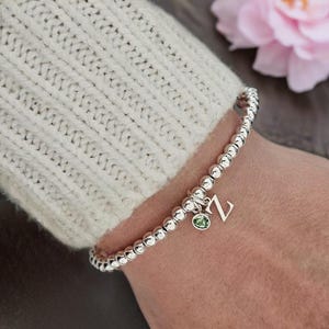 May include: A silver beaded stretch bracelet featuring a small, dangling silver letter Z charm and a single round, light green gemstone bead. The bracelet is composed of polished metallic spheres strung together in a continuous circular design.