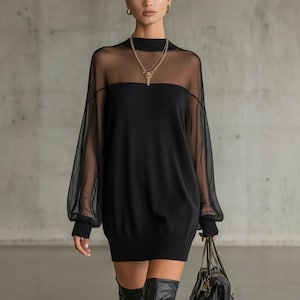 May include: Black mini dress with sheer long sleeves and a high neckline. The dress features a solid bodice and a sheer yoke. The model is wearing a gold necklace and earrings, and carrying a black handbag. The dress is paired with black knee-high boots.