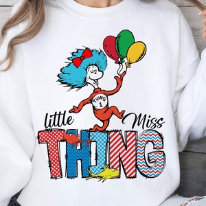 Könnte beinhalten: Weißes Sweatshirt mit einem Cartoon-Charakter mit blauen Haaren, einer roten Schleife und Luftballons. Der Text "little Miss THING" wird in bunten, gemusterten Buchstaben angezeigt. Das Design ist verspielt und skurril.