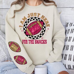 May include: Cream-coloured sweatshirt featuring a football design. The design includes a pink football with gold detailing, set within a black and white chequered circle. Text reads "I AM JUST HERE FOR THE SNACKS". Two footballs are on the left sleeve.