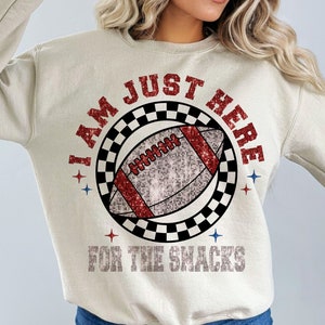 May include: Beige sweatshirt featuring a football graphic. The design includes a football with red and silver glitter details, encircled by a black and white chequered pattern. Text reads "I AM JUST HERE FOR THE SNACKS" in red and silver.