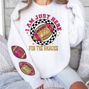 May include: White sweatshirt featuring a football design. The graphic includes a football with pink and gold detailing, and the text "I AM JUST HERE FOR THE SNACKS". Two footballs are also printed on the sleeve.