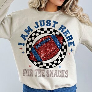 May include: Cream-coloured sweatshirt featuring the text "I AM JUST HERE FOR THE SNACKS". The design includes a football with red and blue glitter, surrounded by a black and white chequered circle and blue stars.