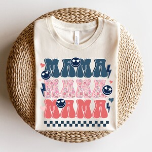 May include: A cream-colored t-shirt with the text "MAMA" in blue and pink retro-style lettering. The letters are decorated with hearts, stars, and lightning bolts.