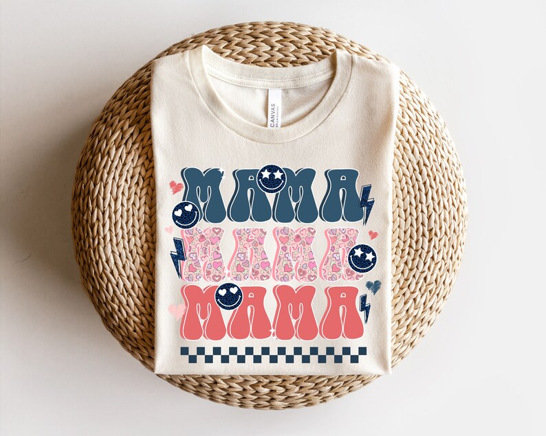 May include: A cream-colored t-shirt with the text "MAMA" in blue and pink retro-style lettering. The letters are decorated with hearts, stars, and lightning bolts.