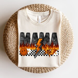 May include: A white t-shirt with the word "MAMA" printed in black with flames and a lightning bolt. The design also includes a smiley face, a butterfly, and a checkered pattern.