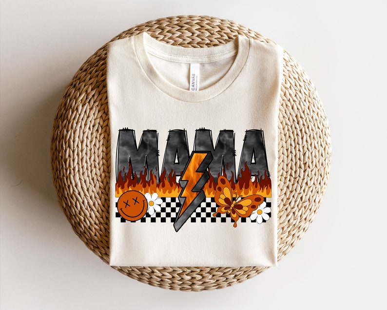 May include: A white t-shirt with the word "MAMA" printed in black with flames and a lightning bolt. The design also includes a smiley face, a butterfly, and a checkered pattern.