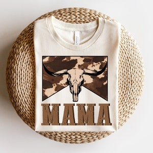 May include: A white t-shirt with a brown and white cow skull graphic and the word "MAMA" in brown text.
