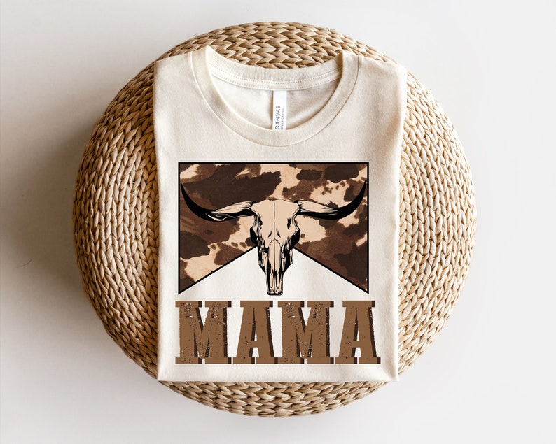 May include: A white t-shirt with a brown and white cow skull graphic and the word "MAMA" in brown text.