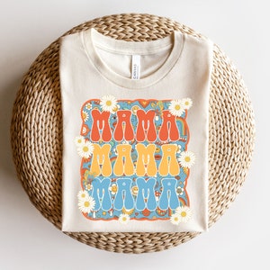May include: A beige t-shirt with a retro-style graphic design featuring the word "Mama" repeated three times in different colors. The graphic is surrounded by daisies and other psychedelic patterns.