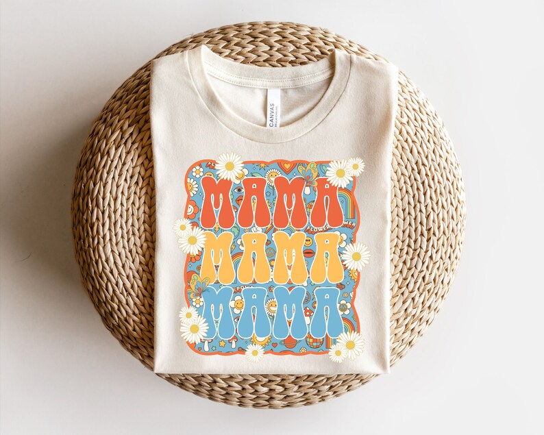 May include: A beige t-shirt with a retro-style graphic design featuring the word "Mama" repeated three times in different colors. The graphic is surrounded by daisies and other psychedelic patterns.