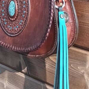 May include: A brown leather saddle bag featuring intricate embossed circular patterns, braided leather trim, and a central oval turquoise stone inlay. The bag is accented with a long, bright turquoise leather fringe tassel attached to a silver-tone ring.