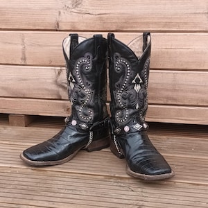 May include: A pair of black leather cowboy boots featuring a square toe, a textured crocodile-embossed finish on the foot, and intricate white embroidery on the shaft. The boots include a removable harness strap with silver studs and pink accents.