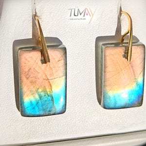 Puede incluir: Un par de pendientes rectangulares de labradorita con tonos azules y melocotón iridiscentes, suspendidos de ganchos de metal dorado. Las piedras muestran fracturas internas naturales y una superficie pulida. El texto TUMAY CREATIONS es visible.