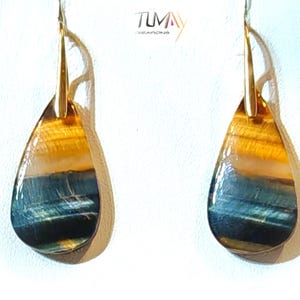 Peut inclure: Paire de boucles d'oreilles en forme de goutte d'eau avec des crochets dorés. La pierre polie présente des bandes bleues, dorées et marron. Les boucles d'oreilles sont exposées sur une surface blanche. Le nom de la marque "TUMAY CREATIONS" est visible.
