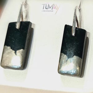 May include: A pair of rectangular earrings with a dark, possibly black, upper section and a silver-colored lower section. The earrings have a hook-style closure. The brand name "TUMAY CREATIONS" is visible in the background.