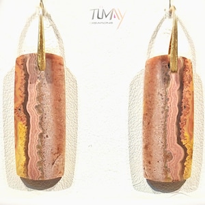 May include: A pair of rectangular earrings with a gold-colored top and a natural stone design. The stone features bands of pink, brown, and yellow hues. The earrings are displayed on a white surface.