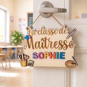 Peut inclure: Une plaque de porte en bois clair suspendue à une poignée argentée, portant l'inscription La classe de Maîtresse SOPHIE en lettres sombres et colorées. Elle est décorée de découpes en bois représentant un crayon, une calculatrice et une palette.