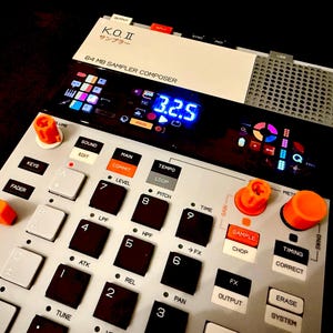 Puede incluir: Un sampler y compositor de música digital, con una interfaz blanca y gris. Cuenta con numerosos botones, perillas naranjas y una pantalla digital que muestra "3.2.5". El texto "K.O. II" y "64 MB SAMPLER COMPOSER" es visible.