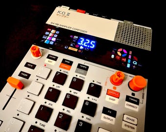 Peut inclure: Un échantillonneur et compositeur de musique numérique avec une interface blanche et grise. Il comporte de nombreux boutons, des boutons orange et un écran numérique affichant "3.2.5". Le texte "K.O. II" et "64 MB SAMPLER COMPOSER" est visible.