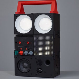 Peut inclure: Un haut-parleur modulaire noir et rouge avec une poignée, deux haut-parleurs blancs, un écran numérique et un grand haut-parleur en bas.