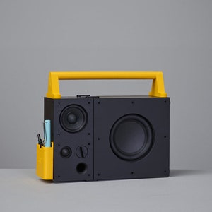 May include: A black speaker with a yellow handle and pen holder. The speaker has a large circular woofer and smaller speakers. A light blue pen and a black pen are in the yellow pen holder.