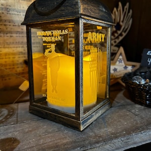 May include: A decorative lantern with a black and gold finish. The glass panels feature etched designs of a soldier saluting and an American flag. The lantern contains a glowing candle, and text that reads "PAT NICHOLAS DORMAN" and "OF ARMY".
