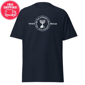 Puede incluir: Una camiseta azul marino con un diseño circular blanco que presenta una menorá y texto en hebreo. El texto dice "Mossad" y "El escudo de Israel".